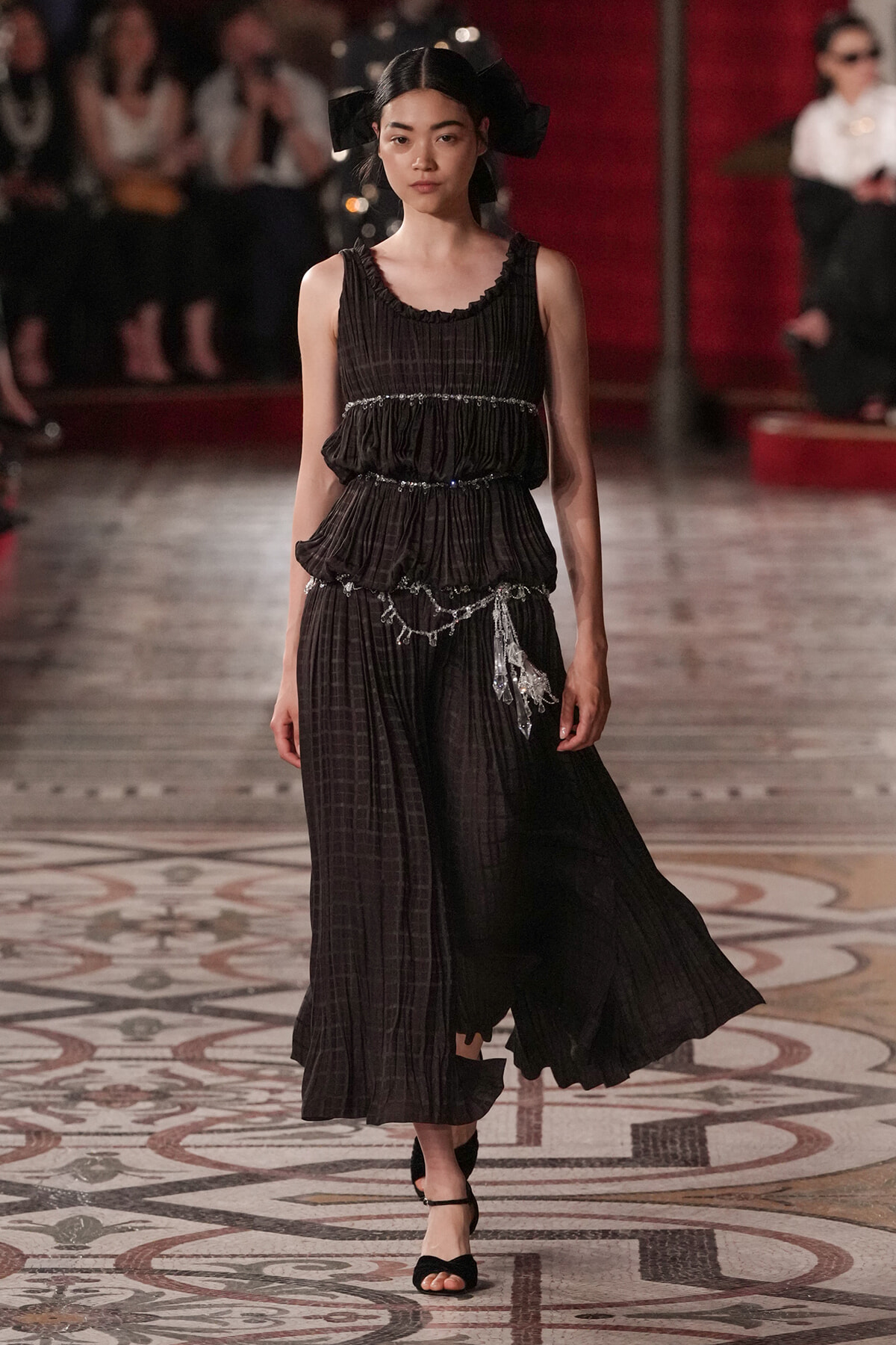 Model walks a runway in a black pleated dress with layered ruffles and silver chain belt, on a patterned floor.