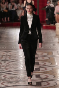 Model walking the runway in a black velvet suit with a white high‑neck blouse and a jeweled bib, her hair tied with oversized black bows.
