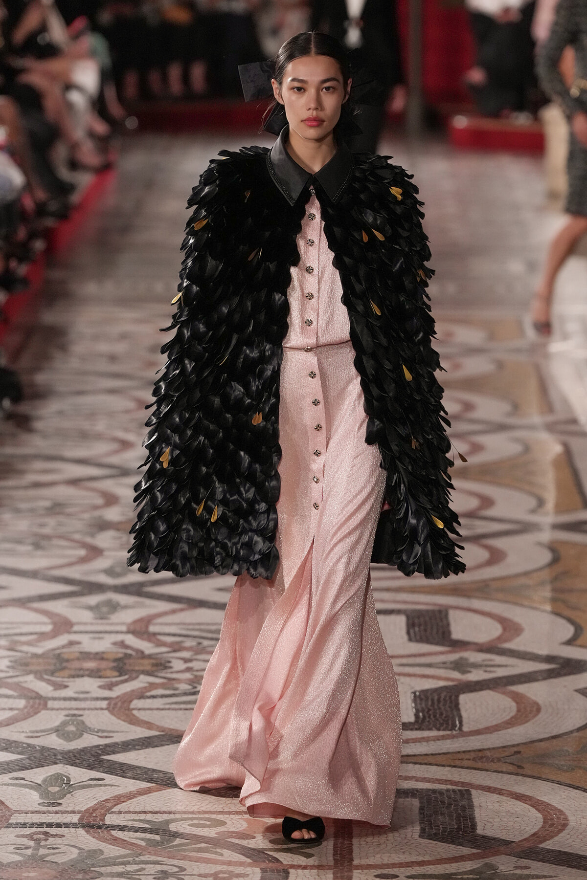 Model walking runway in a pale pink, buttoned gown with a dramatic black feathered cape.