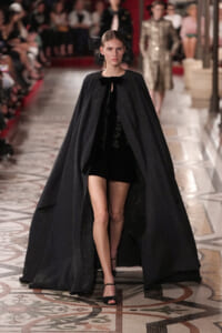 Runway model in a long black textured cape over a short black dress with a bow at the neck, walking the catwalk in black sandals.