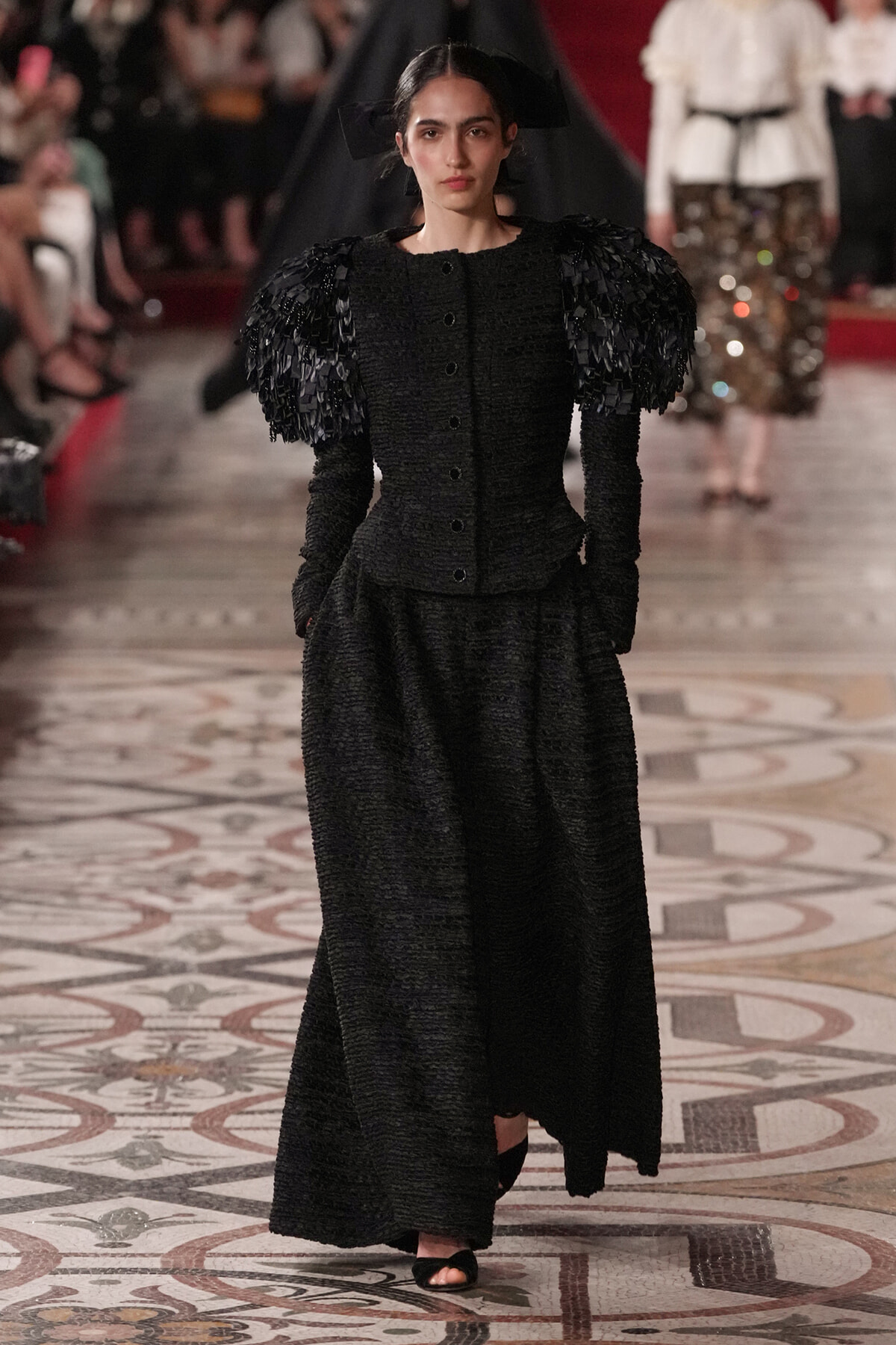 Model on a runway wearing a long black textured gown with large sculptural shoulder poufs.