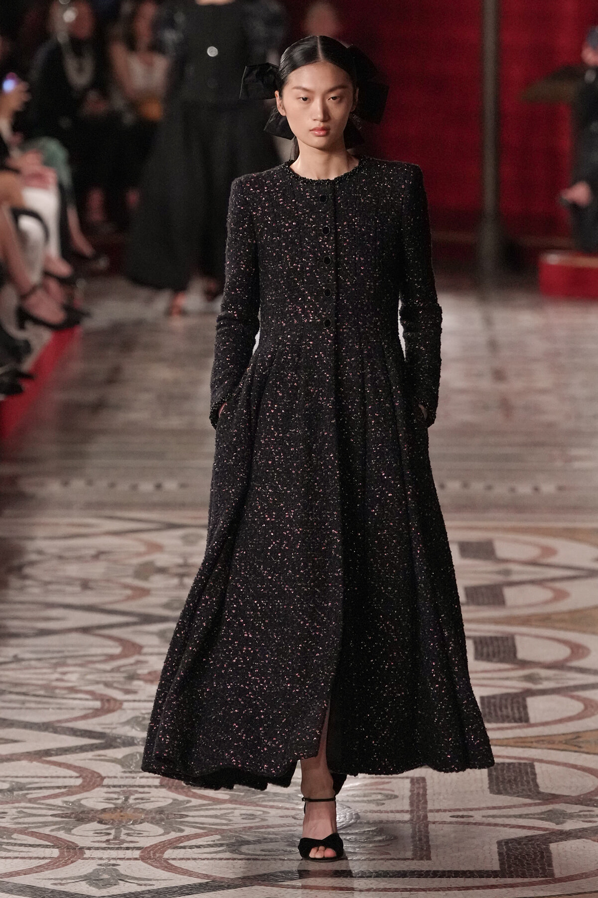 Model walking the runway in a full-length black speckled dress with buttoned bodice and pockets, wearing a black headpiece.