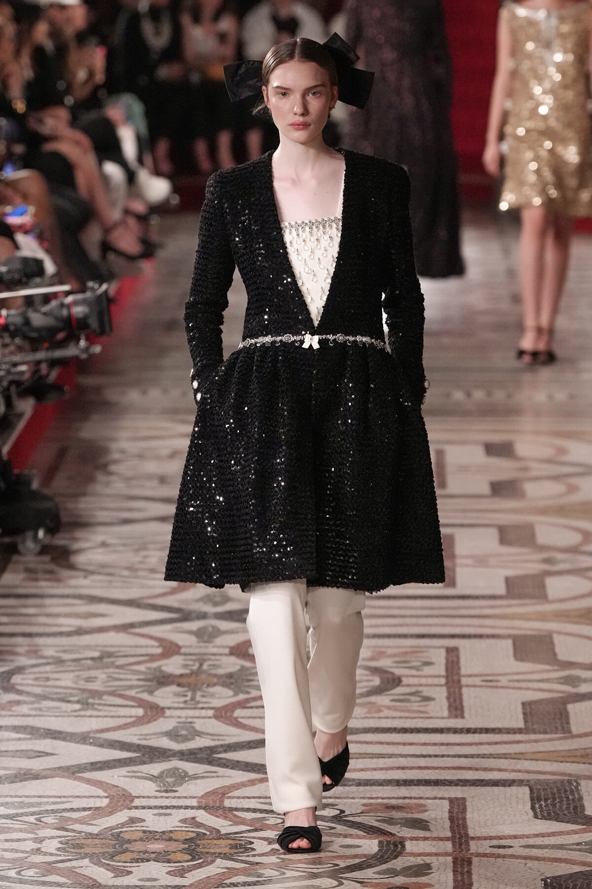 Model on a runway wearing a black sequined coat-dress over a white embellished top, with a slim belt and white pants, plus a large black bow headpiece.