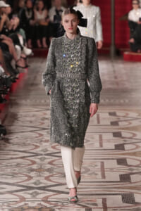 Model walks the runway in a silver textured coat with colorful bead embellishments and a large black bow headpiece.