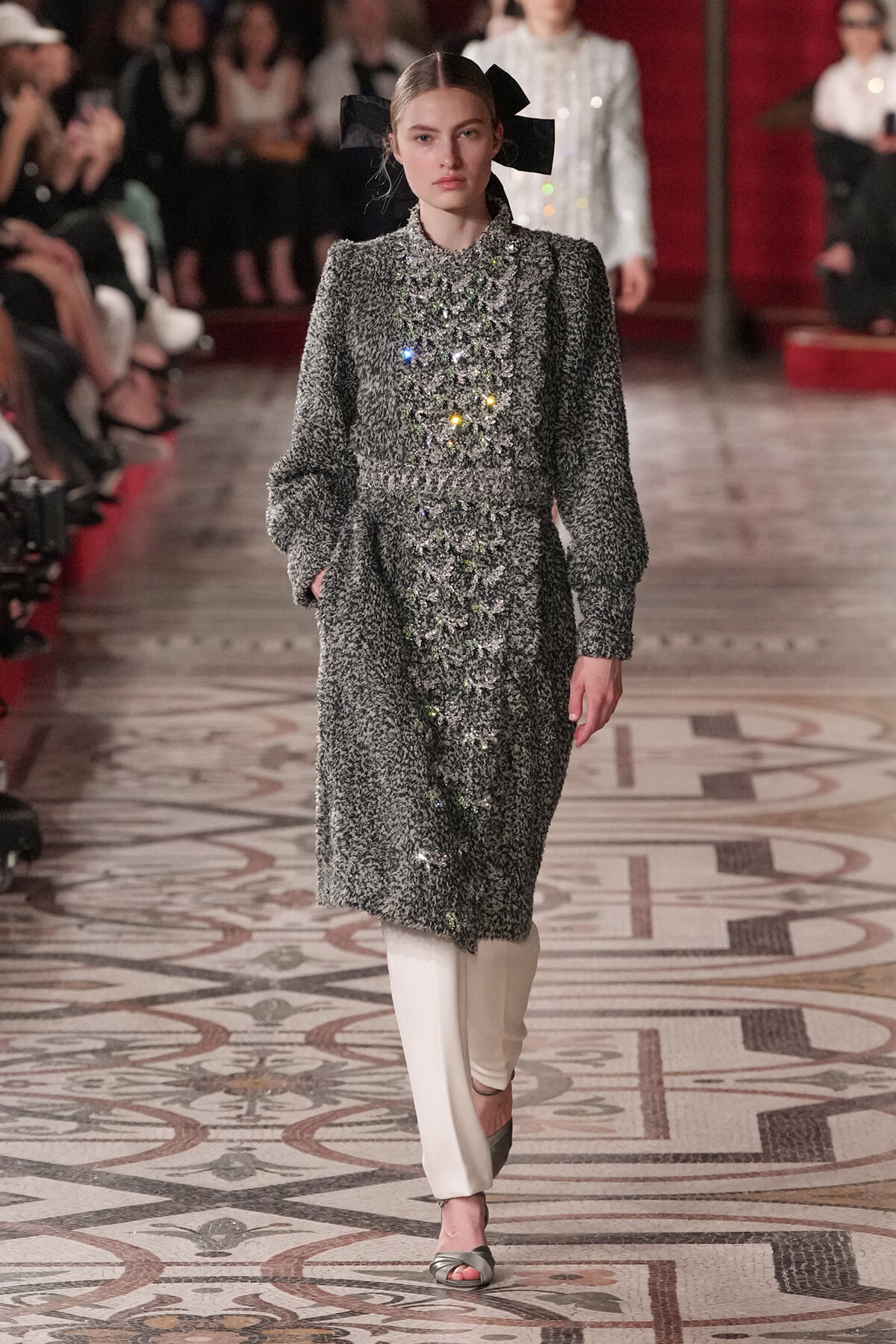 Model walks the runway in a silver textured coat with colorful bead embellishments and a large black bow headpiece.