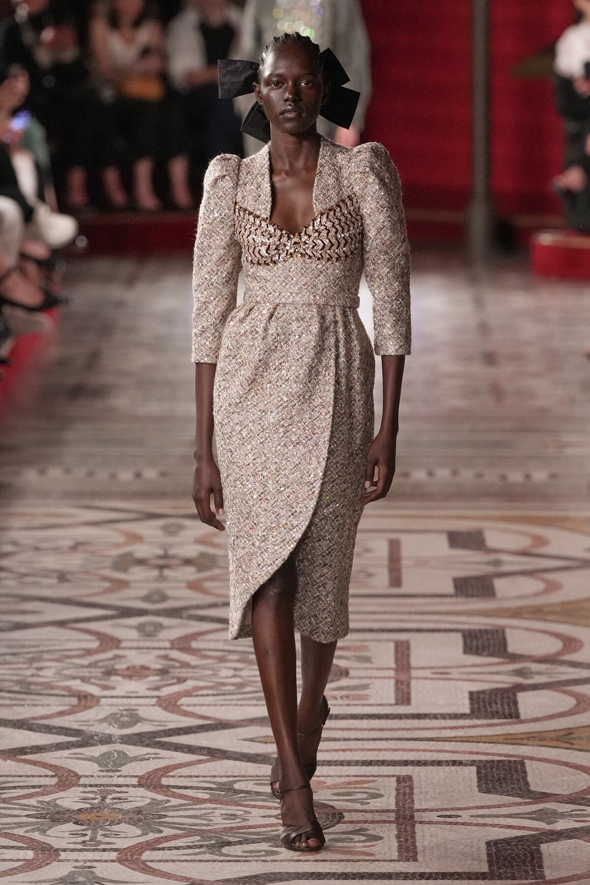 Model on a runway wearing a sparkling champagne dress with sculpted shoulders and a wrap skirt.