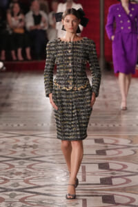 Model on a runway wearing a black-and-white tweed dress with gold buttons and a matching belt, plus a large black bow headpiece and gray ankle-strap heels.