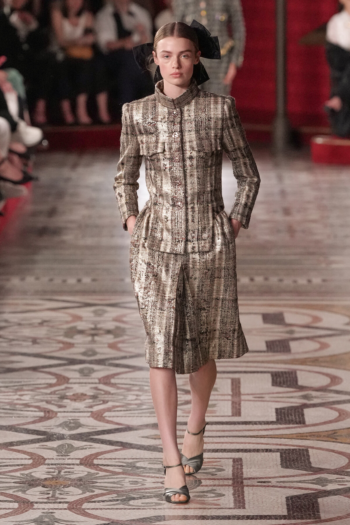 Female model on a runway wearing a silver tweed tailored jacket with a matching knee-length skirt, black bow headpiece, and gray sandals.
