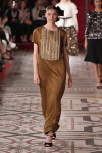 Model walks the runway in a shimmering gold gown with a vertical beaded panel, plus a large black bow headpiece and black sandals.