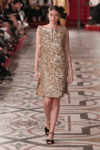 Model walking a runway in a gold sequined sleeveless dress with a large black bow headpiece.