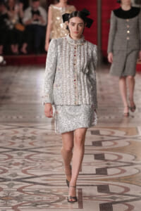 Model walking a runway in a silver embellished tweed suit with a high collar and bold black bow in her hair