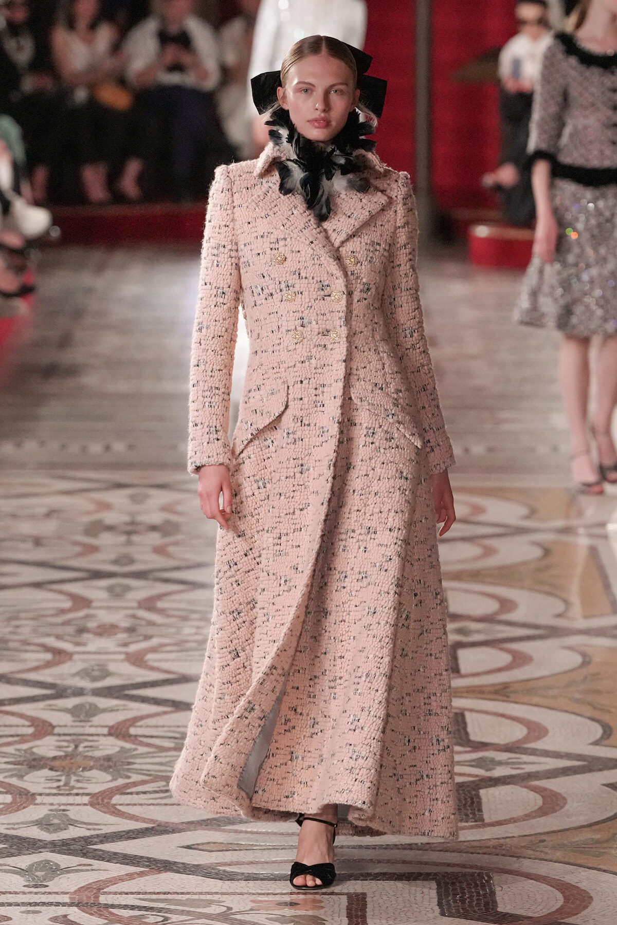 Model walking runway in a pale pink tweed long coat with double-breasted gold buttons and a matching skirt, accessorized with a black feather scarf and headpiece.