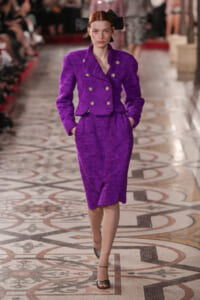 Model on a fashion runway wearing a purple textured double-breasted suit with gold buttons and a black bow headpiece.