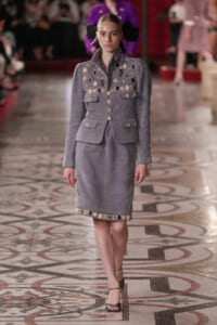Model walking a fashion show in a gray embellished suit with gold buttons and a matching skirt; large black bow headpiece in back.