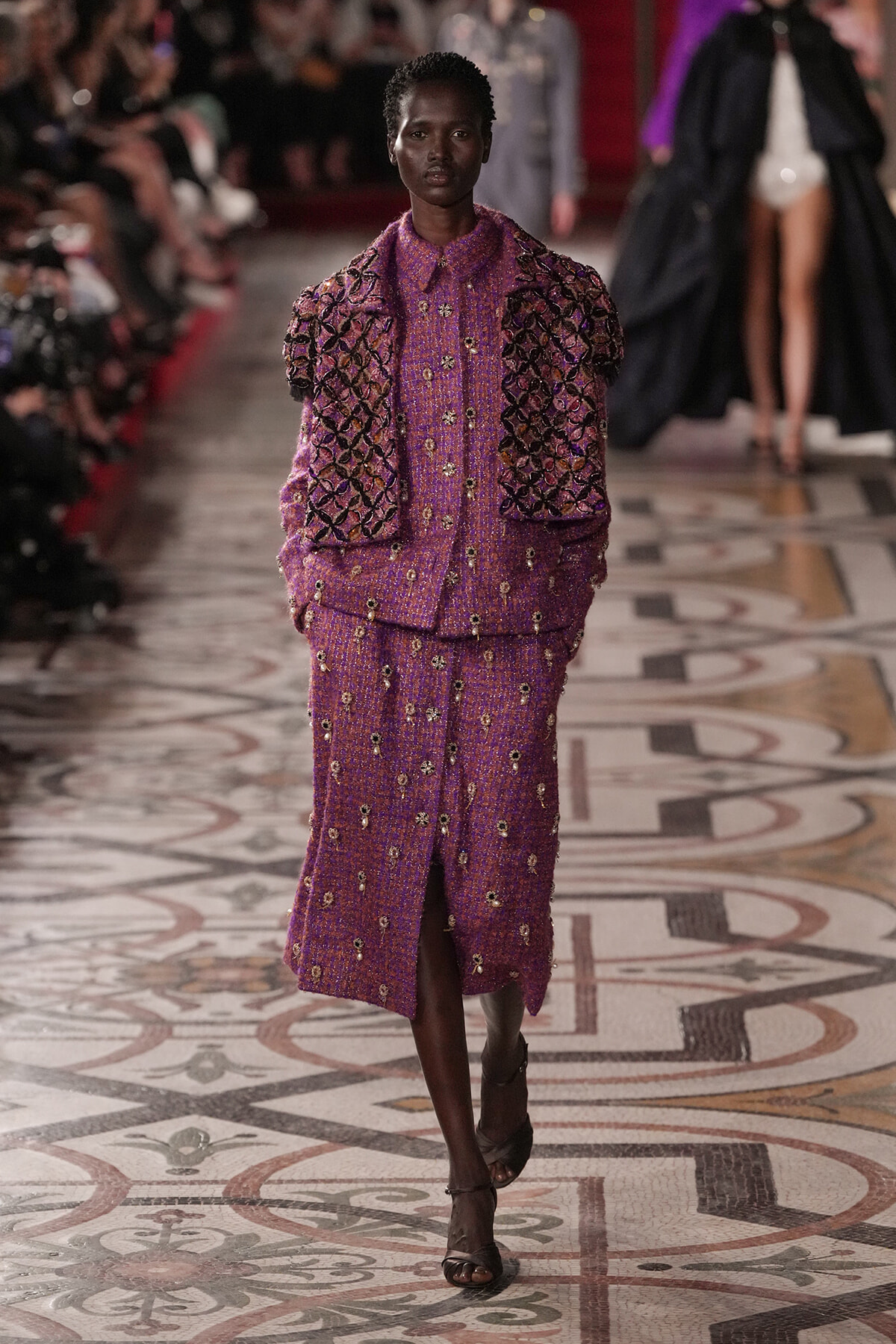 Model walks the runway in a purple beaded two-piece suit with puffed short sleeves and matching long skirt, during a fashion show.