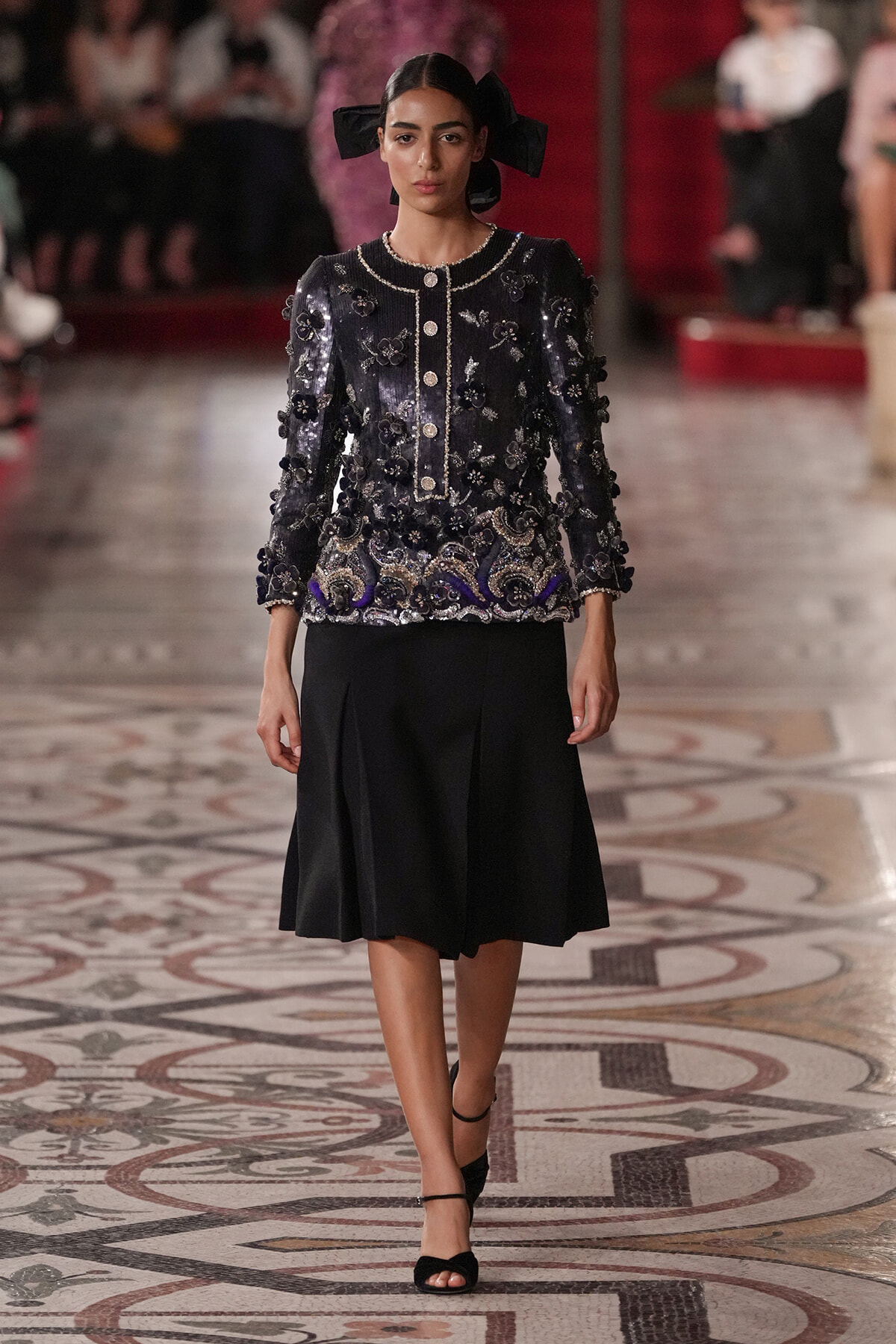 Model walks a runway in a navy, heavily embellished jacket with floral appliqués and a black knee-length skirt, accessorized with a large black bow in hair.