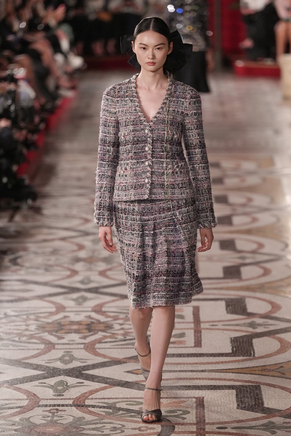 Model walking a fashion runway in a textured gray tweed suit with matching jacket and skirt, black hair accessories in a bow.
