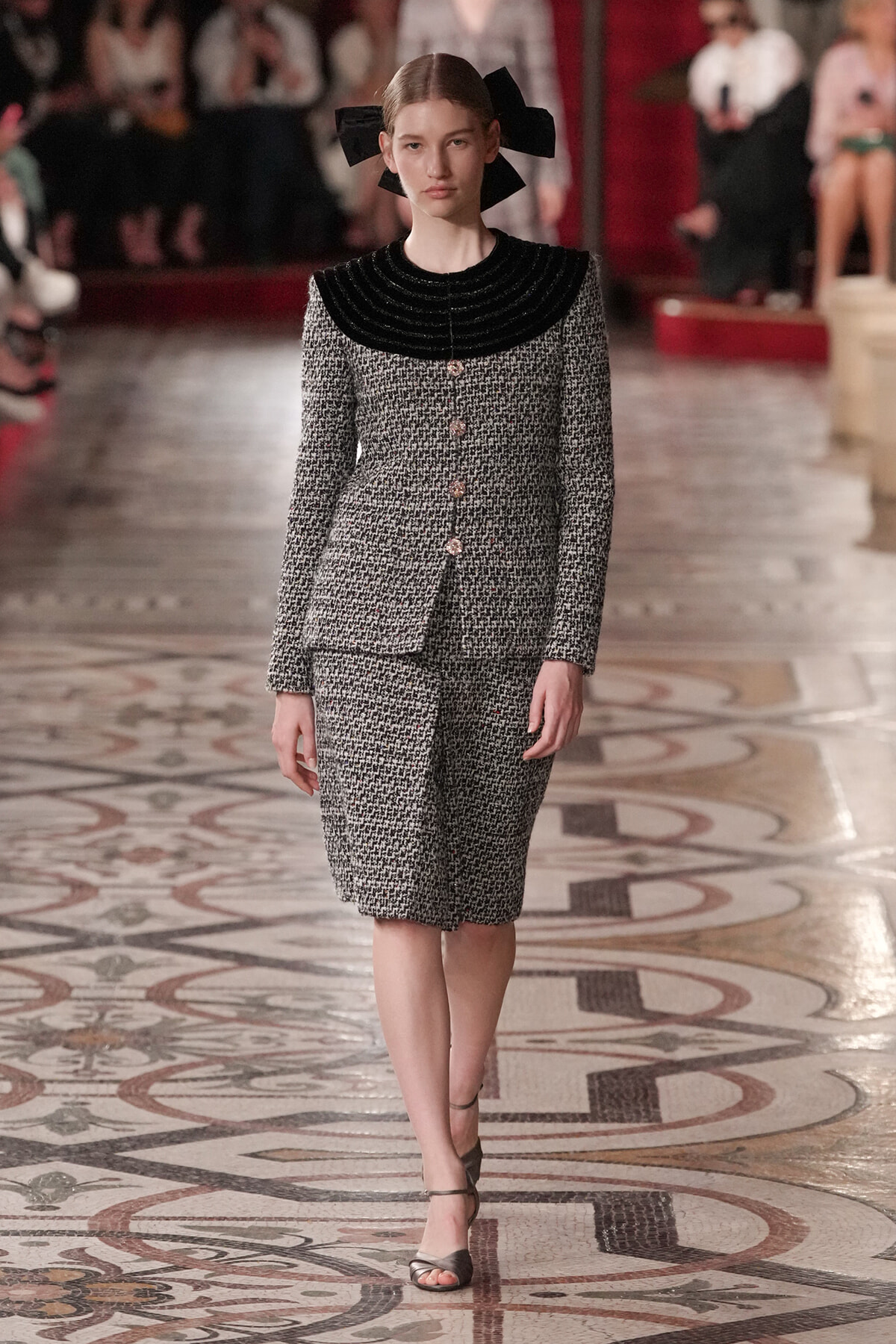 Model on a fashion show runway wearing a textured gray tweed suit with a black circular collar and pink buttons, plus a large black bow in her hair.