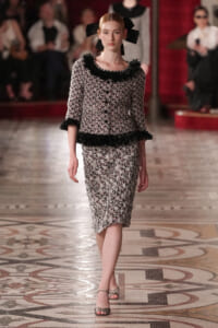 Model walking a fashion runway in a black-and-white tweed suit with lace trim and a large black bow in her hair.