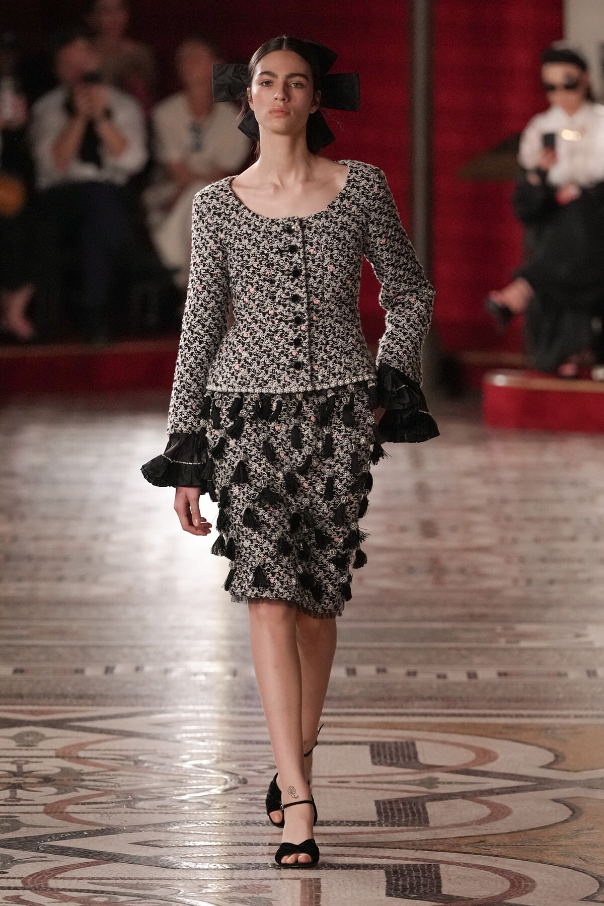 Model walking a fashion runway in a black-and-white floral dress with ruffled hems and a large black bow headpiece.