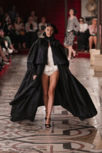 Model walks the runway in a dramatic black cape with a ruffled high collar over a white lace bodysuit, opening to reveal legs.
