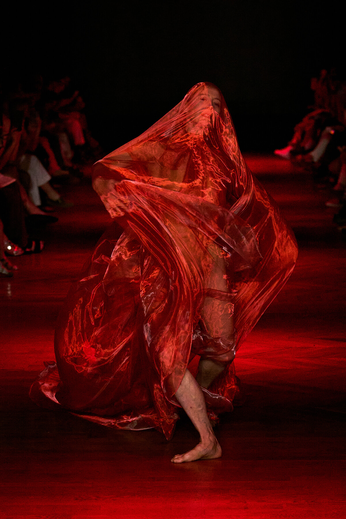 Dancer wrapped in red translucent fabric on a dark stage, barefoot, mid-performance.