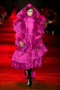 Model on a runway wearing a voluminous magenta ruffled gown with hooded cape and black-and-white boots