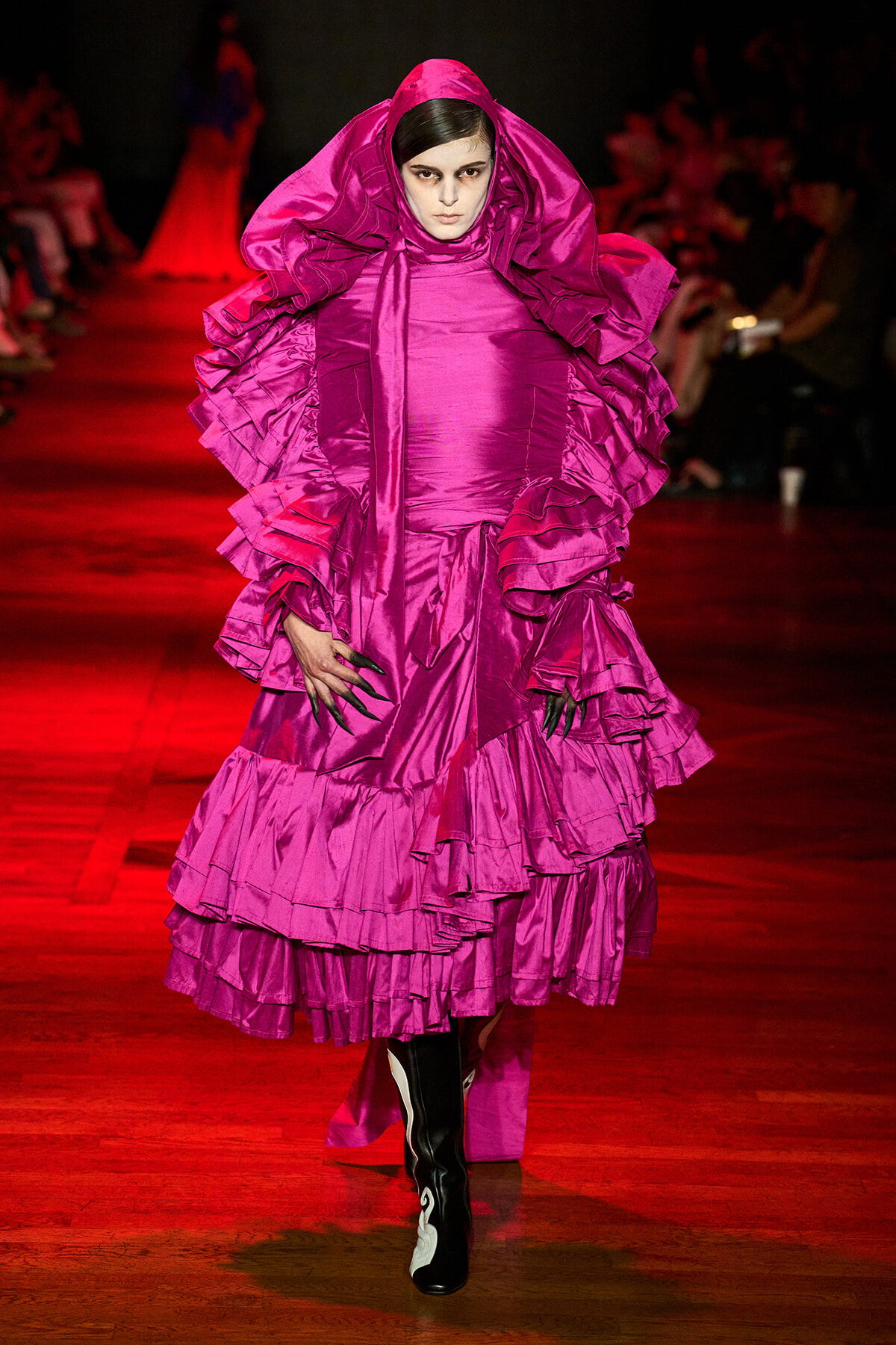 Model on a runway wearing a voluminous magenta ruffled gown with hooded cape and black-and-white boots