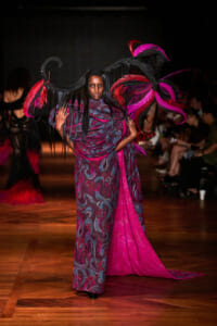 Model on a runway wearing a flowing purple-pink patterned dress with oversized feathered wings in black and magenta, dramatic pose.
