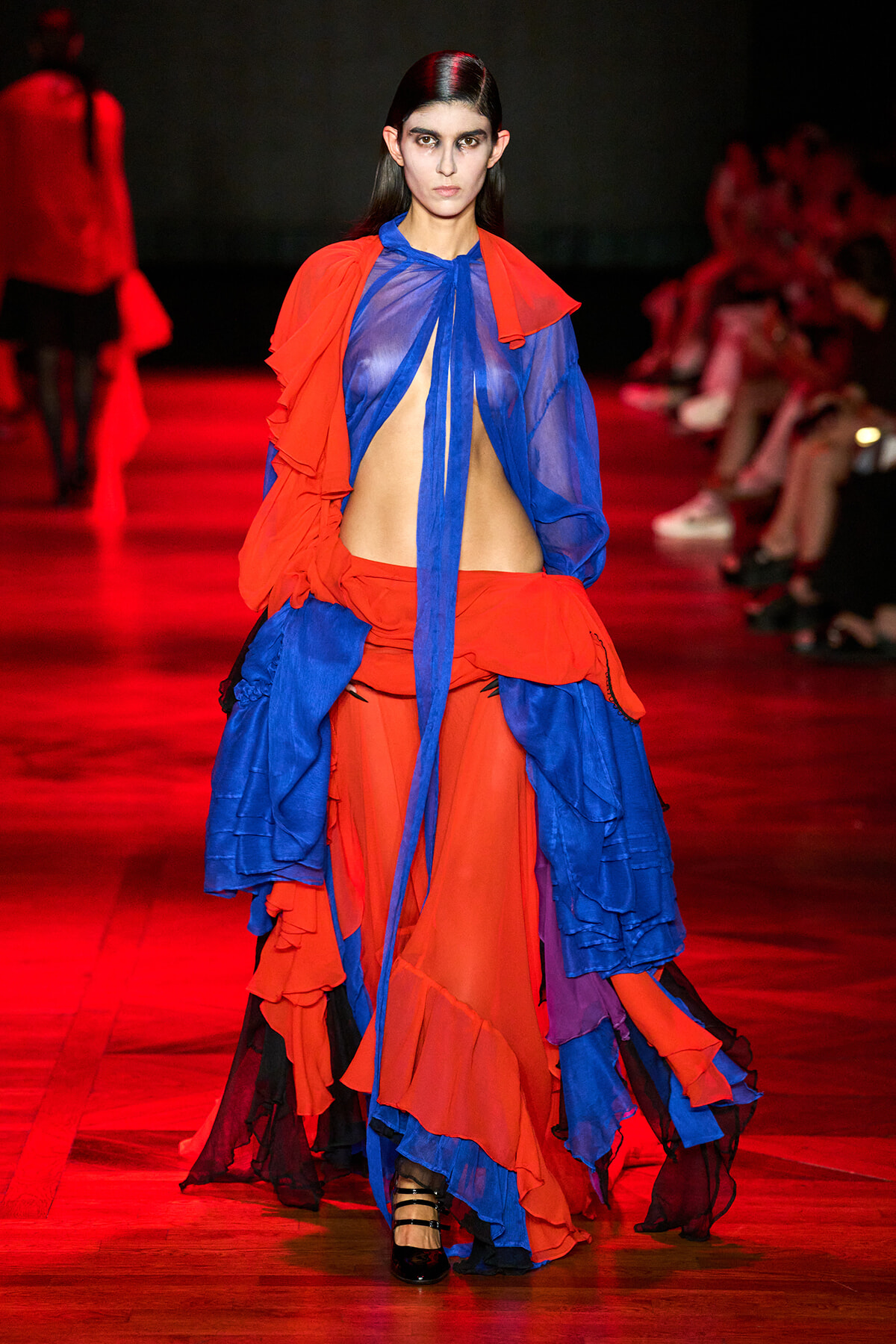 Model on runway wearing a red and blue sheer, ruffled gown with midriff exposed and a long blue neck tie.