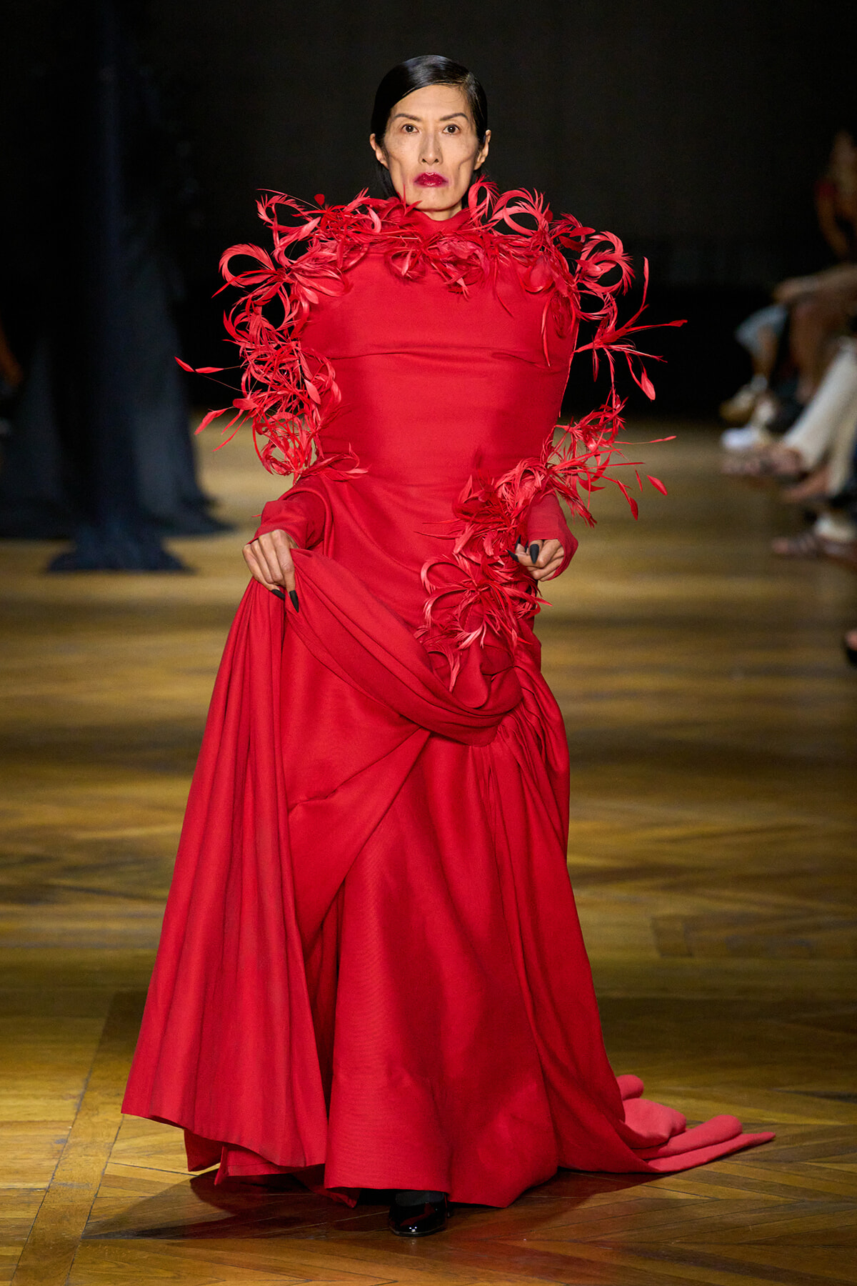 Model on a runway wearing a floor-length red gown with sculptural feathered ruffles along the shoulders and sleeves.