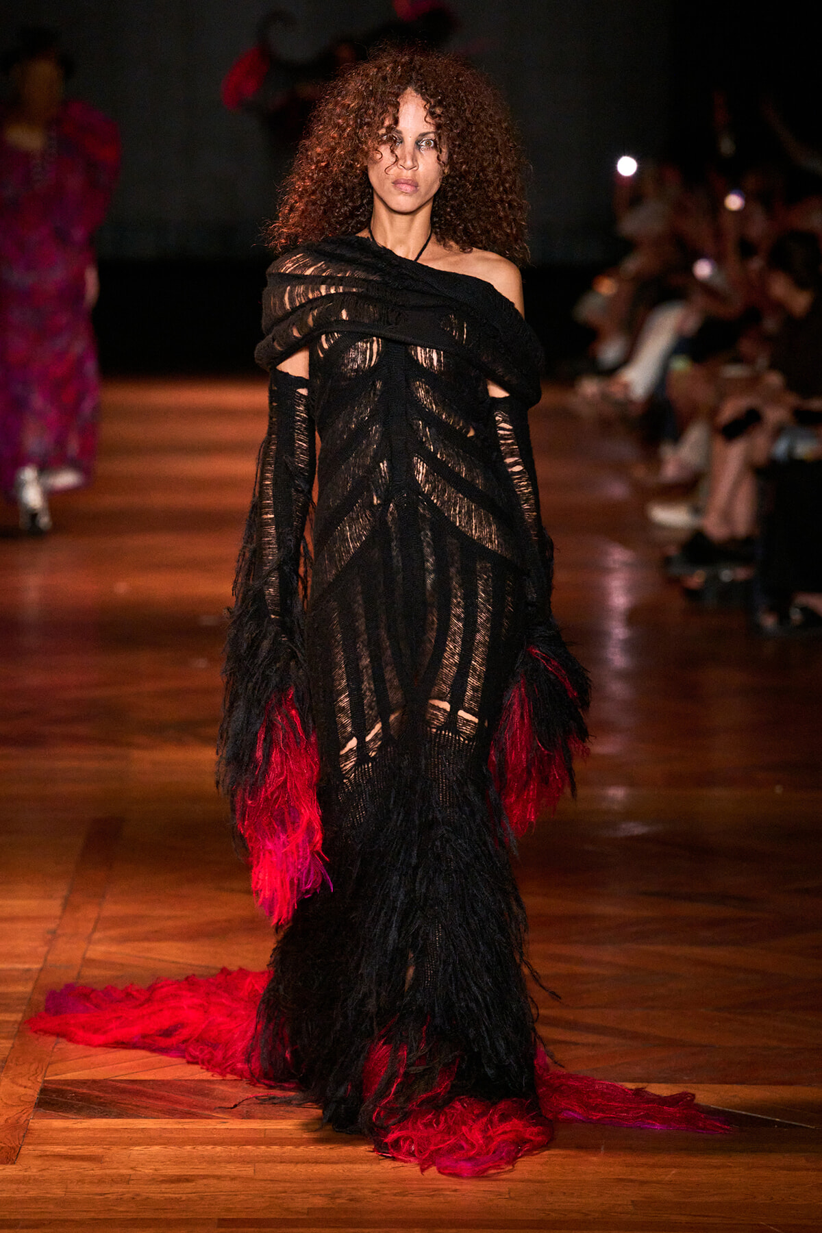 Model walks the runway in a black off-shoulder sheer gown with red and black feather trim cascading to the floor
