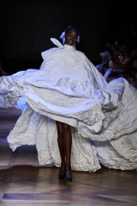 Model walks a fashion runway in a dramatic, oversized pale blue‑gray crinkled gown with billowing folds sweeping the floor.