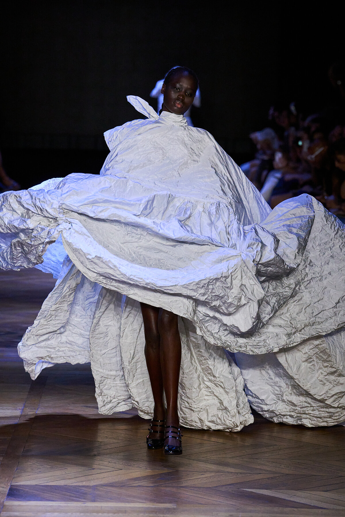 Model walks a fashion runway in a dramatic, oversized pale blue‑gray crinkled gown with billowing folds sweeping the floor.