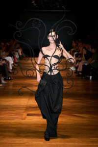 Model walks a runway in an avant-garde black costume with spiraling, wire-like wings over a strapless top and long skirt.