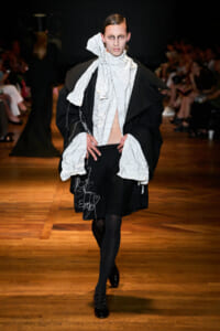 Model on a runway wearing an oversized black cloak with crinkled white scarf and top, mid-stride during a fashion show.