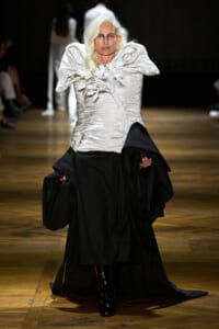 Model on a runway wearing a white sculpted top with floral embellishments and a long black skirt on wooden floorboards.