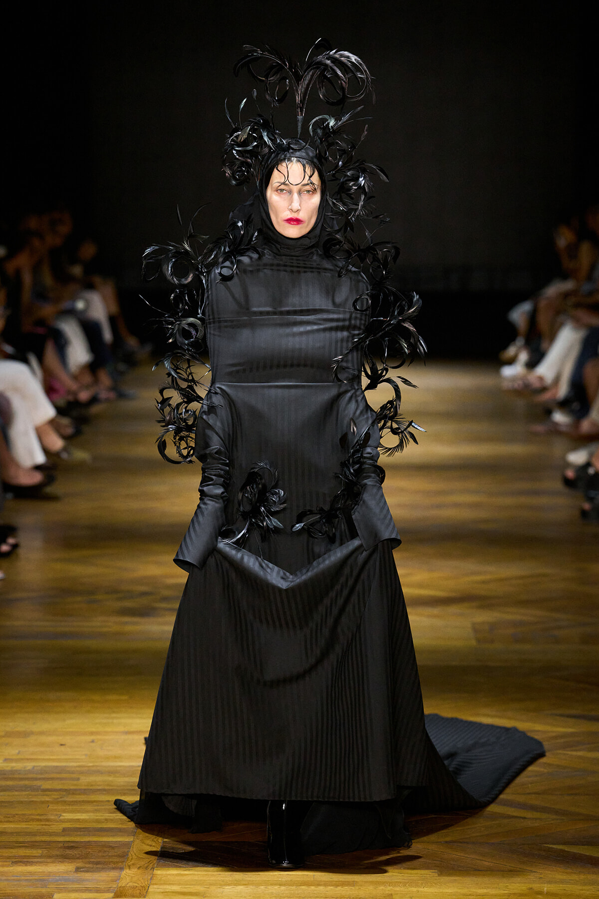 Model walks the runway in a black, pleated gown with an elaborate feather headdress and feather accents along the sleeves.