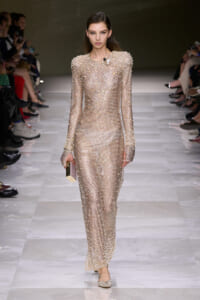 Model on a runway wearing a sparkling, floor-length champagne beaded dress with long sleeves and a chunky pearl necklace, carrying a small clutch.