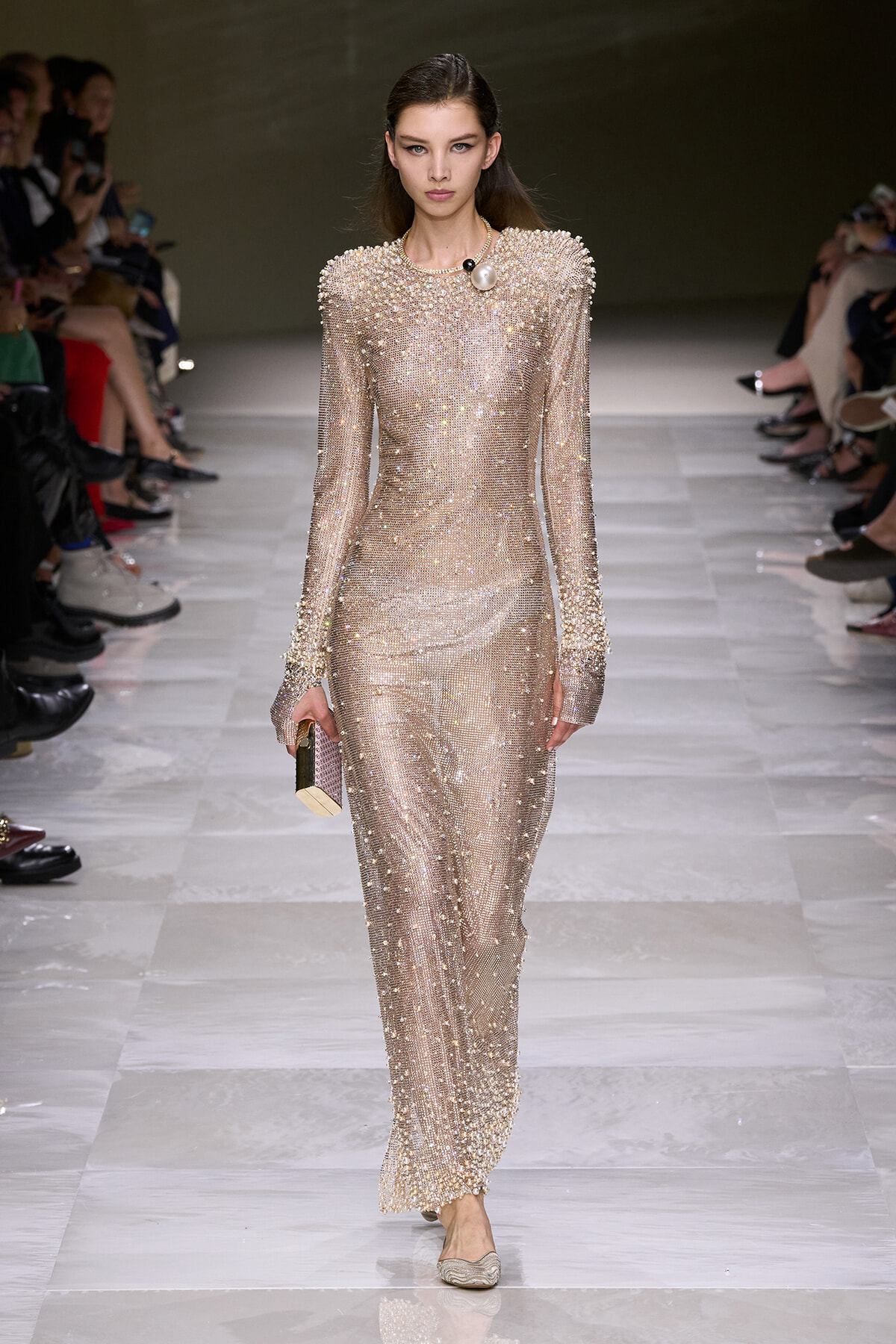Model on a runway wearing a sparkling, floor-length champagne beaded dress with long sleeves and a chunky pearl necklace, carrying a small clutch.