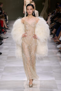 Model walks the runway in a champagne beaded sheer gown with a large white feather cape and chandelier earrings.