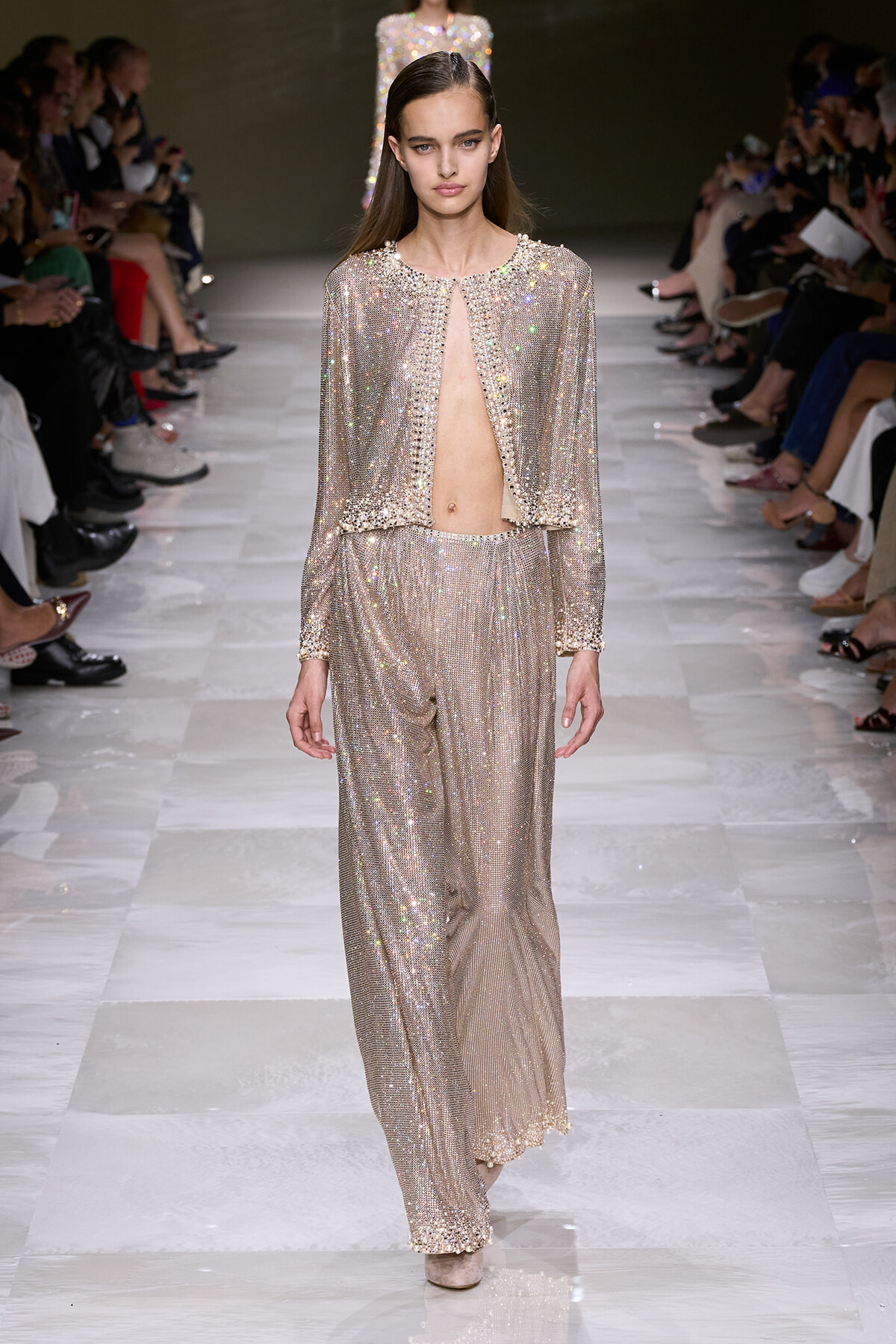 Model walks the runway in a glittering champagne-colored sequin set with a cropped open-front jacket revealing midriff.