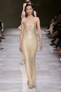 Model walking a runway in a strapless gold beaded gown with a cutout at the waist, on a glossy catwalk.