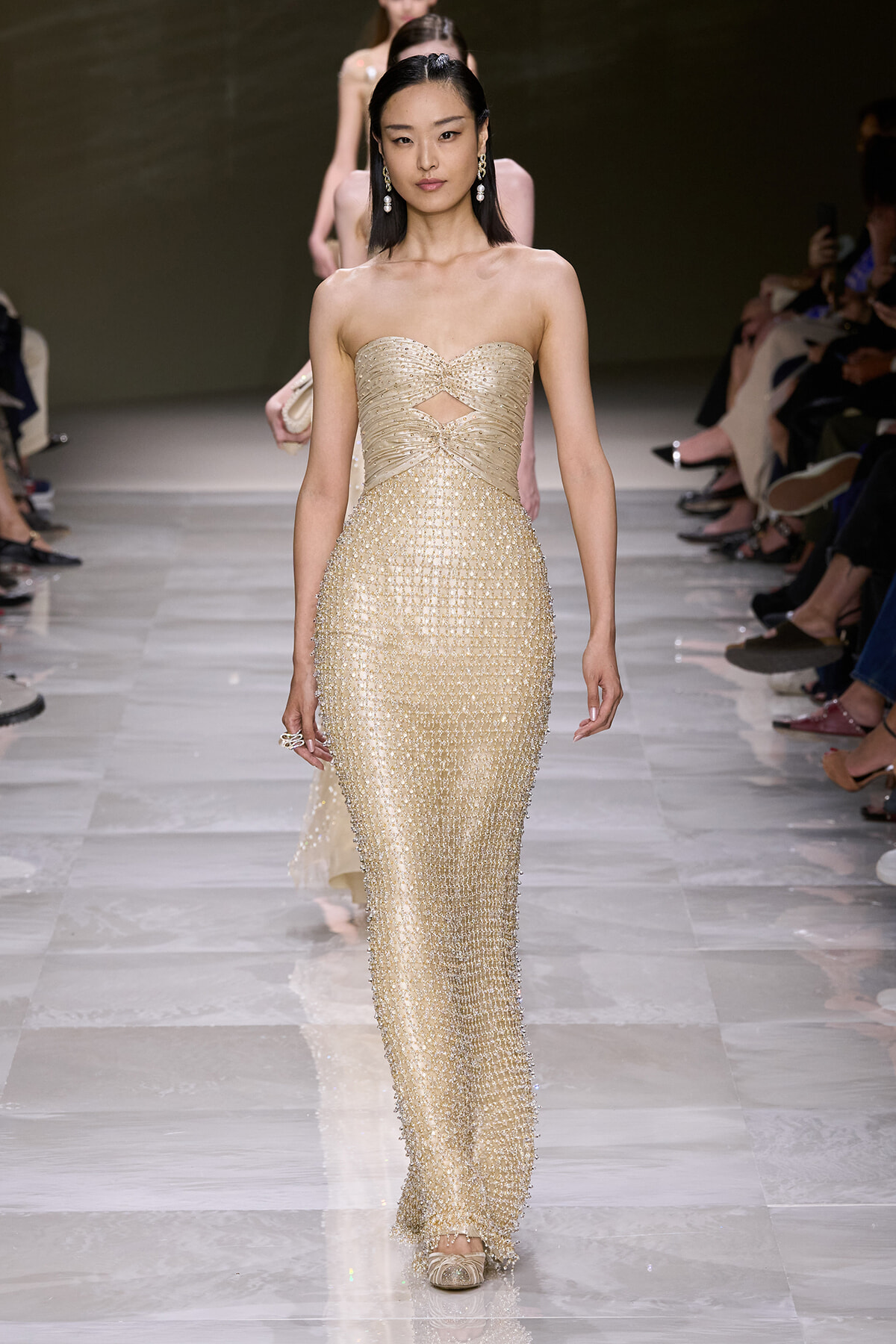 Model walking a runway in a strapless gold beaded gown with a cutout at the waist, on a glossy catwalk.