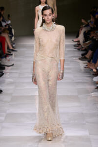 Model walking a runway in a champagne-beige sheer beaded top and full-length embellished lace skirt, adorned with large pearl accessories and cuffs