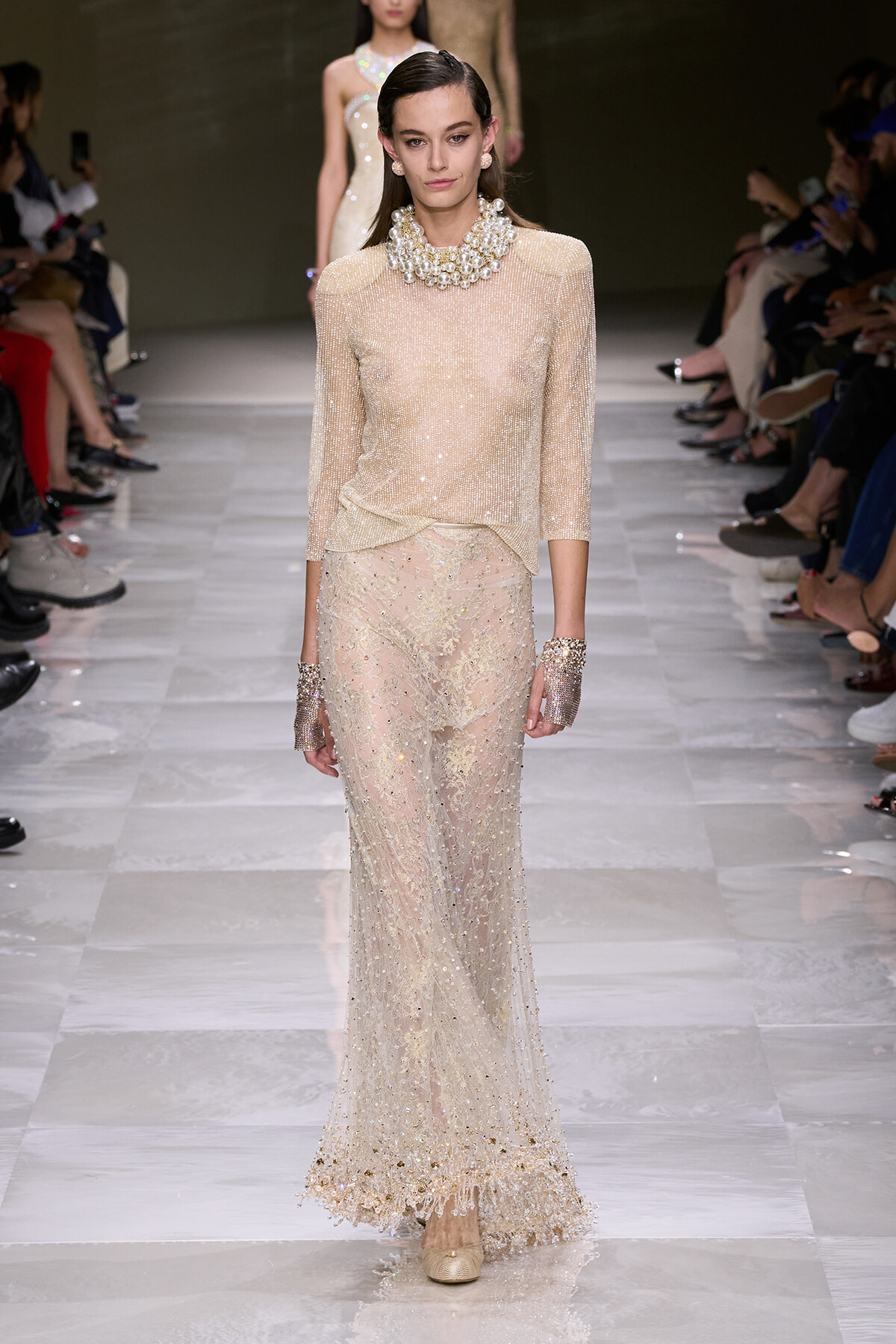 Model walking a runway in a champagne-beige sheer beaded top and full-length embellished lace skirt, adorned with large pearl accessories and cuffs