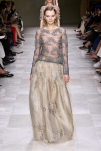 Model walking the runway in a sheer beaded long-sleeve top paired with a champagne floral-embroidered, floor-length skirt.