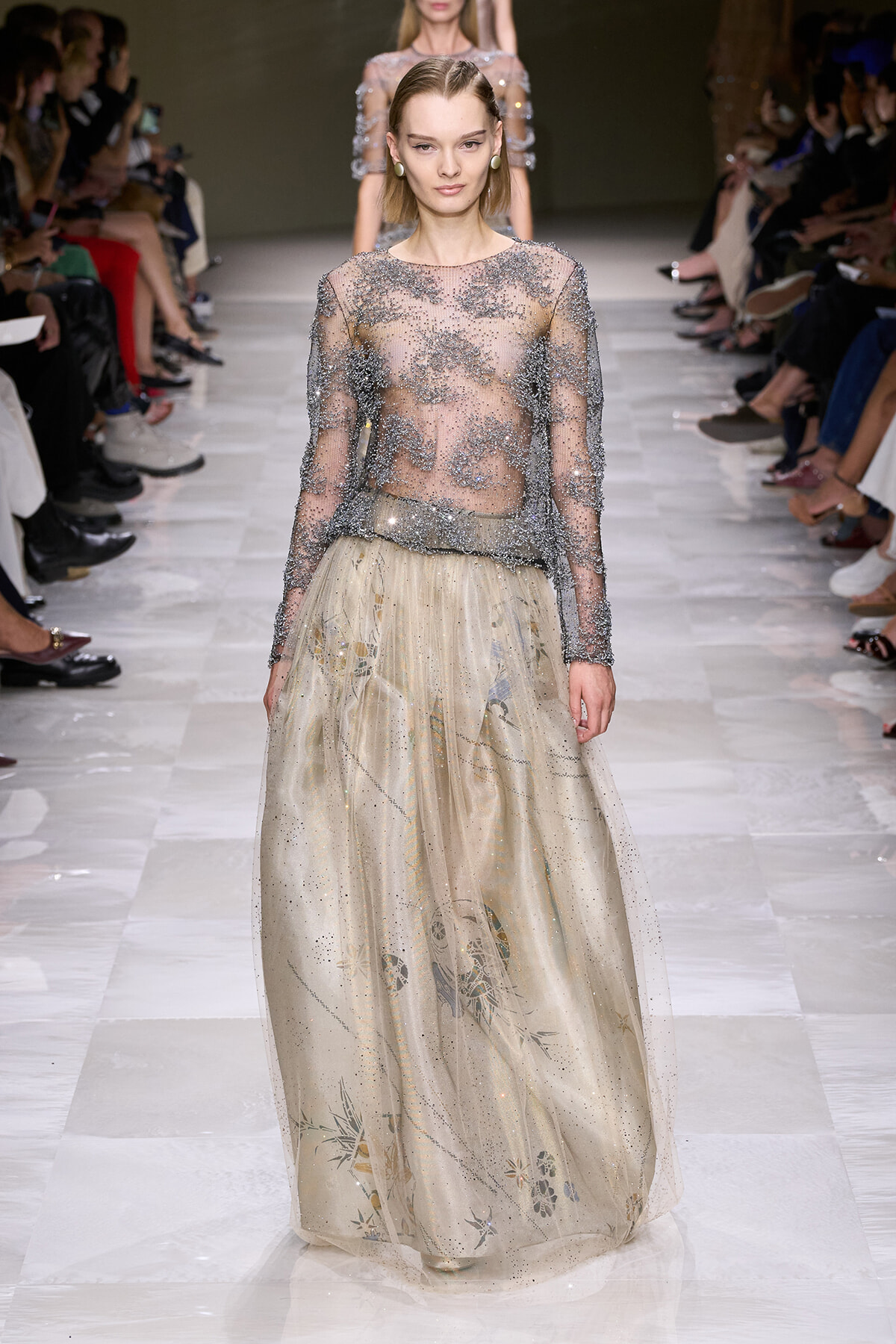 Model walking the runway in a sheer beaded long-sleeve top paired with a champagne floral-embroidered, floor-length skirt.