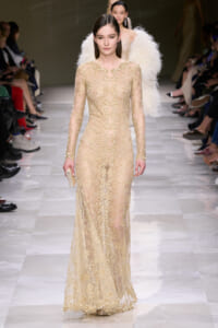 Model walks a long, nude lace gown with gold bead embroidery down a fashion runway; she holds a small beige clutch beside a sheer, embellished silhouette.