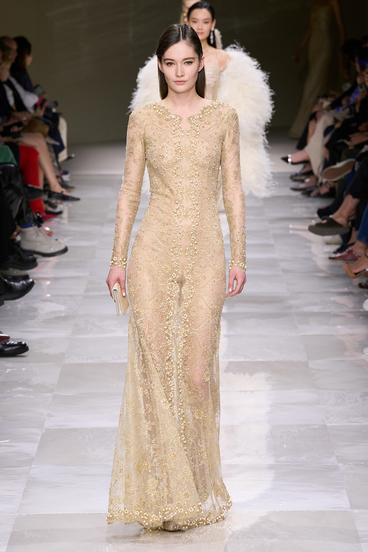 Model walks a long, nude lace gown with gold bead embroidery down a fashion runway; she holds a small beige clutch beside a sheer, embellished silhouette.